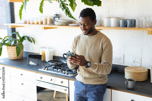Happy african american man leaning on countertop in kitchen, using smartphone