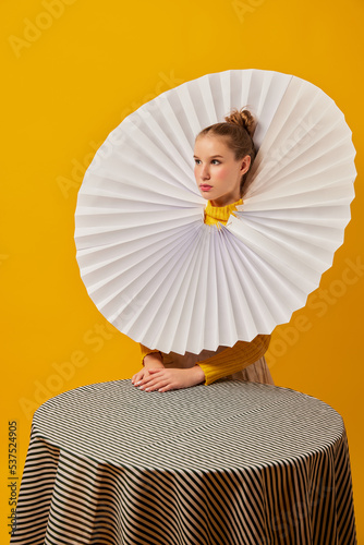 Dinner party. Young indifferent girl in giant jabot collar or neckwear and yellow tights isolated over yellow background. Contemporary art  weird beauty  fashion