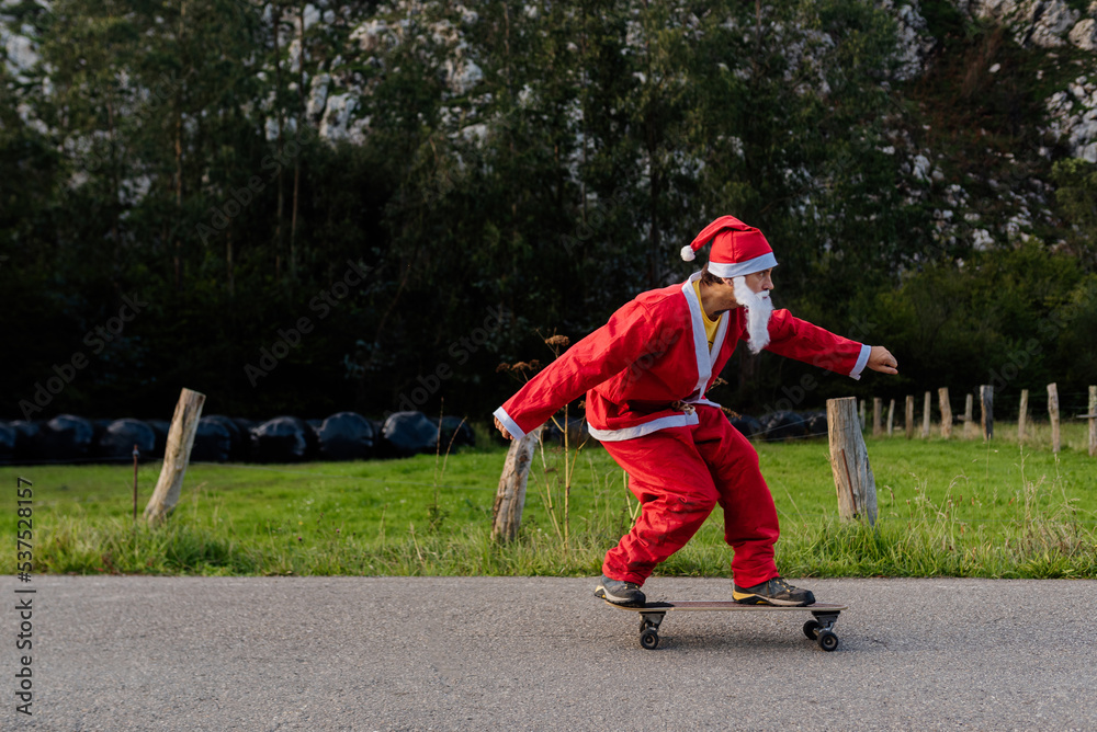 Obraz premium caucasian man dressed as santa claus skating on a longboard skateboard on a country road. christmas and sport concept.