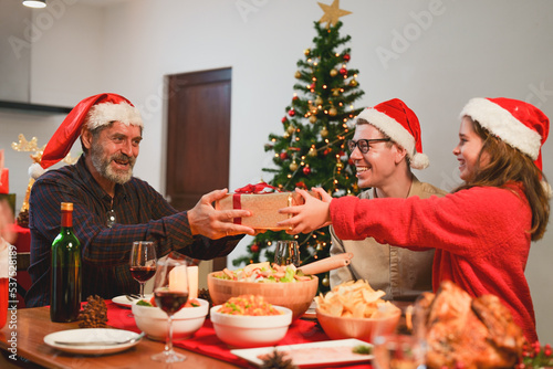 Happy family celebrating Christmas dinner at home. Granddaughter with gift box given to grandfather wearing Santa hats.Merry Christmas or New Year s eve concept