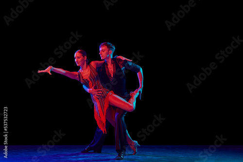 Full-length portrait of young beautiful man and woman dancing ballroom dance isolated over dark background in neon light. Concept of art, beauty, grace, action, emotions. © master1305