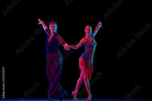 Stylish ballroom dancers couple in gorgeous outfits dancing in sensual pose on dark background in neon light. Concept of art, music, dance, emotions.