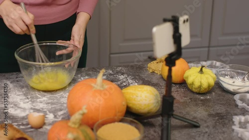 unrecognizable blogger record video live stream cooking pumpkin pie in kitchen smartphone camera on tripod stand table. make dough beat eggs with metal whisk in transparent bowl. thanksgiving day meal photo