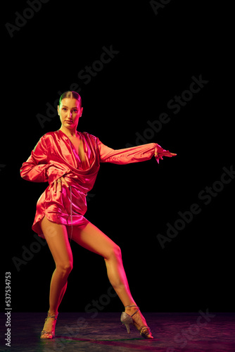 Solo. Portrait of young graceful flexible woman dancing ballroom dance without partner isolated on dark background in neon light.
