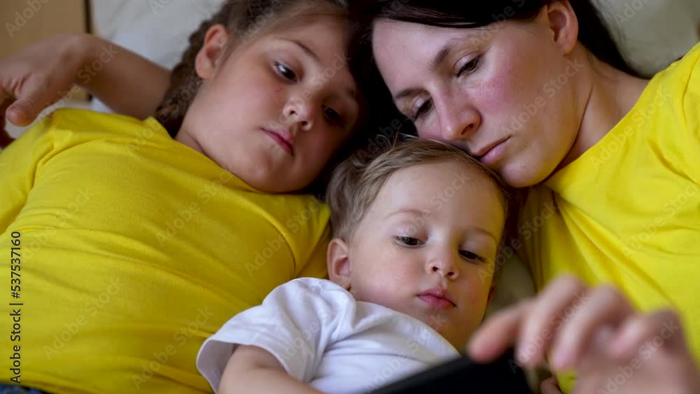 Happy family concept. Mom, daughter and son are play on bed. Child watching video on smartphone. Family leisure at home. Boy and girl with mom are watching movie on smartphone. Home education concept Happy family concept. Mom, daughter and son are play on bed. Child watching video on smartphone. Family leisure at home. Boy and girl with mom are watching movie on smartphone. Home education concept