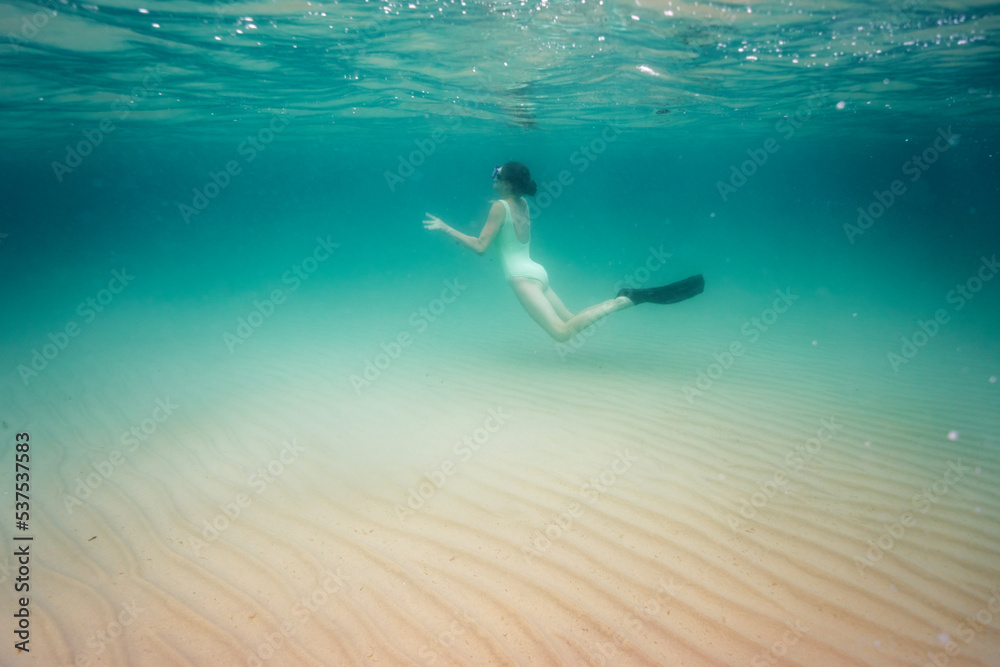 A teenage girl in an underwater mask dives to the sandy bottom at a shallow depth in the sea while holding her breath.