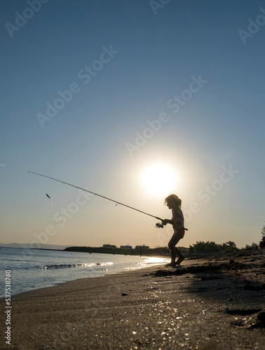 little girl is fishing with a fishing rod in the sea, under the sun. Silhouette. © Yasin
