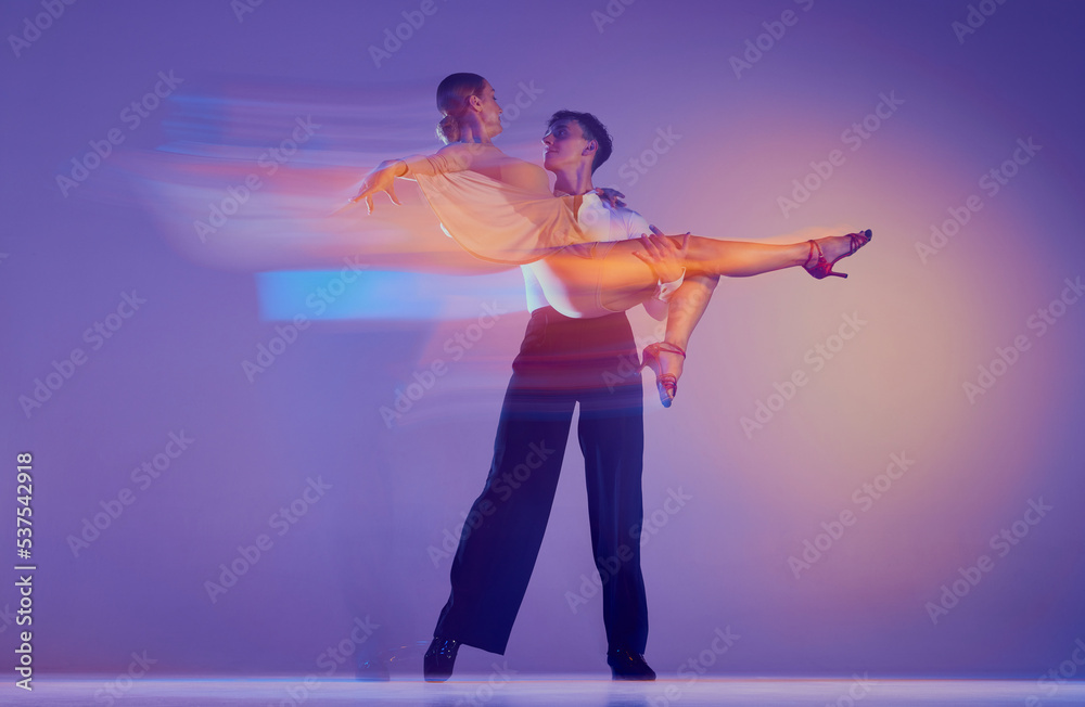 Art in motion. Flexible adorable ballroom dancers in stage attires dancing isolated on purple background in neon mixed light. Music, dance, emotions Art in motion. Flexible adorable ballroom dancers in stage attires dancing isolated on purple background in neon mixed light. Music, dance, emotions