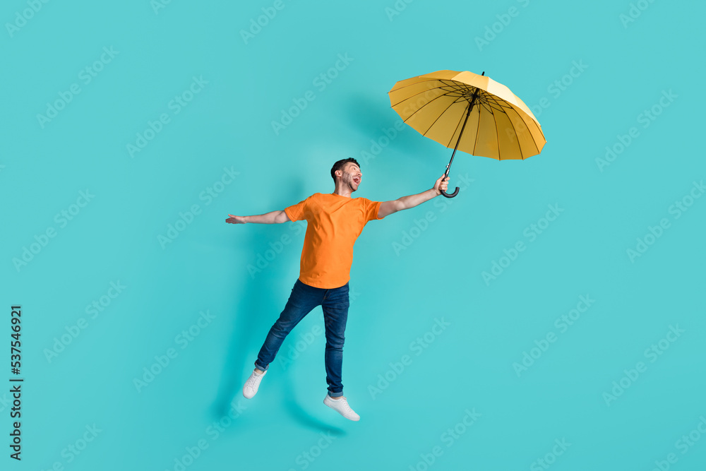 Full length photo of positive man stylish orange clothes yellow parasol flying empty space isolated on aquamarine color background Full length photo of positive man stylish orange clothes yellow parasol flying empty space isolated on aquamarine color background