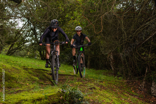 Two female cyclists wearing goggles and helmets riding their bikes down a mountain in the middle of a forest