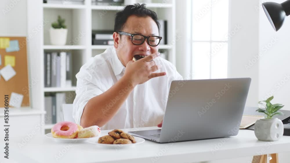 Asian fat man eating donut, sweet, junk food during working with computer laptop, unhealthy eating concept Asian fat man eating donut, sweet, junk food during working with computer laptop, unhealthy eating concept