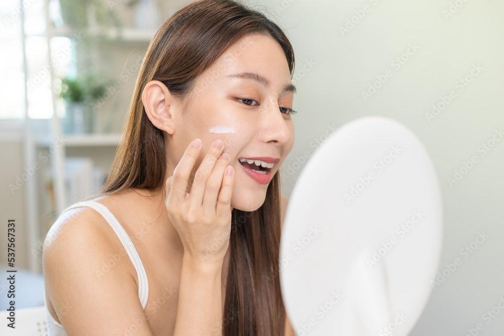 Fresh healthy skin, beautiful smile of asian young woman, girl looking at mirror, applying moisturizer on her face, putting cream treatment before makeup cosmetic routine at home. Facial Beauty. Fresh healthy skin, beautiful smile of asian young woman, girl looking at mirror, applying moisturizer on her face, putting cream treatment before makeup cosmetic routine at home. Facial Beauty.