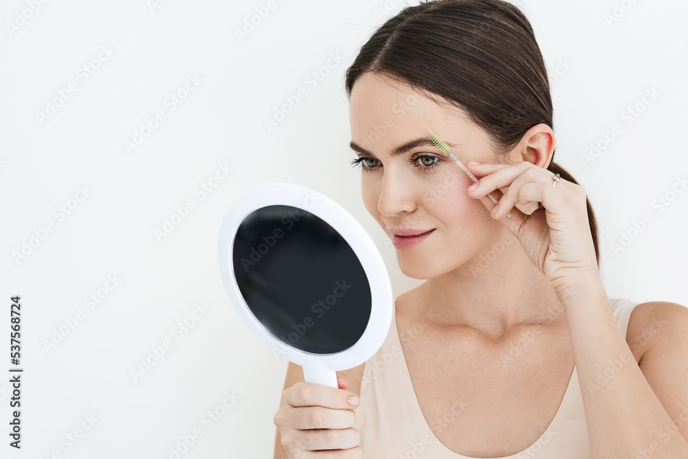 Beautiful woman combing her eyebrows with brush tool while looking into portable mirror, isolated on white. Eyebrow brushing process Beautiful woman combing her eyebrows with brush tool while looking into portable mirror, isolated on white. Eyebrow brushing process
