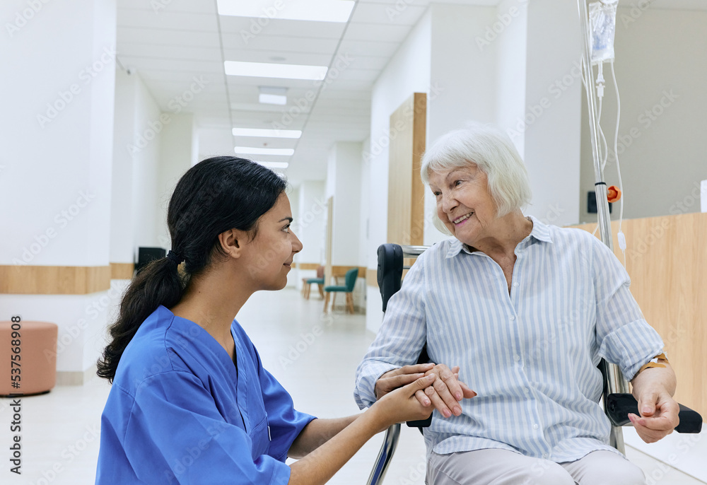 Obraz premium Psychological support from nurse to elderly woman sitting in wheelchair on corridor of medical clinic. Caring for people with disabilities at hospital
