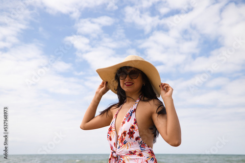 Beautiful young woman posing on the beach with a hat to protect herself from the sun. The woman is enjoying her trip to a paradise beach. Holiday and travel concept. © @skuder_photographer