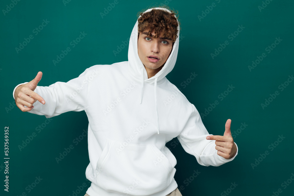 a handsome man stands on a green background in a white hoodie with a hood over his head and points his fingers at his clothes