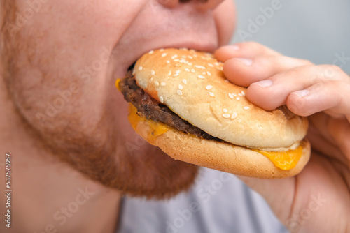 The man bites and chews a double cheeseburger. A man eats fast food. Unhealthy diet, bad habits.