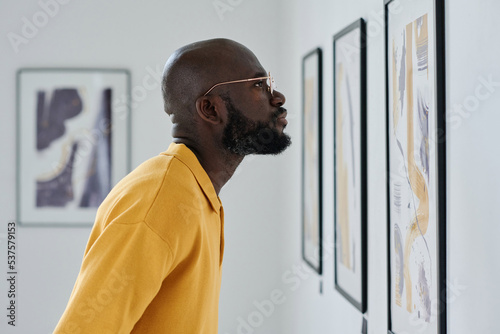 African man in eyeglasses looking at pictures on the wall while visiting art gallery