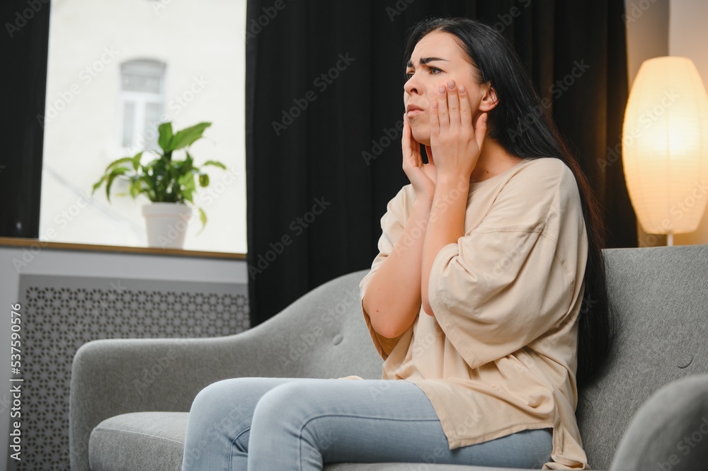 Woman with severe toothache touching her swollen cheek. Young girl feeling discomfort after tooth extraction. Upset lady with inflamed tooth nerve or terrible dental pain sitting on sofa at home.