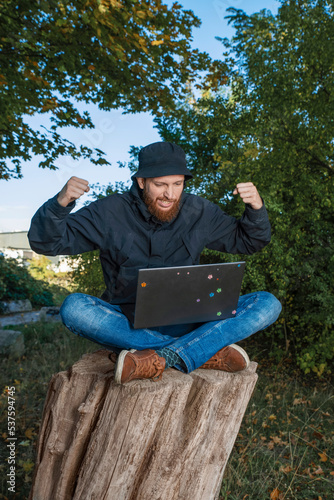 man with a laptop rejoices at the information received, works remotely sitting on a stump in nature, concept