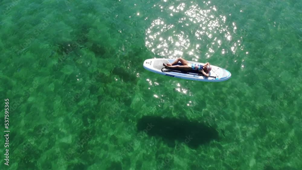 Aerial view from drone: young attractive woman in bikini and sunglasses floating on paddle board on turquoise water