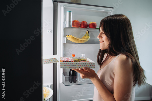 Young Woman Enjoy Eating Donut From Plate Near Refrigerator In Kitchen