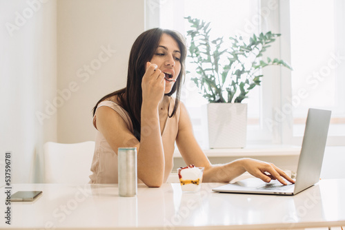 The concept of working at home. Young woman with a laptop in the kitchen, working with pleasure and eating delicious ice cream.