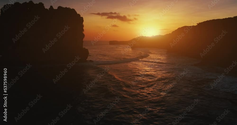 Orange sunset cliff coast silhouette. Ocean bay waves slow motion aerial view. Close up rocky mountain on sea water backdrop. Tropical rock shore at yellow evening sun. Summer travel Bali, Indonesia.