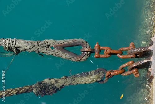 Two ship ropes seen up close, chains and rusty hooks attached to the concrete wall over the turquoise sea of ​​Salvador, Bahia, Brazil.