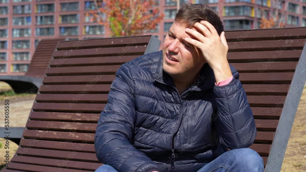 Pensive mature man wearily rubs his forehead while sitting on a wooden chaise longue in a city park. Thoughtful man sitting on bench and looking aside on street. Pensive mature man wearily rubs his forehead while sitting on a wooden chaise longue in a city park. Thoughtful man sitting on bench and looking aside on street.