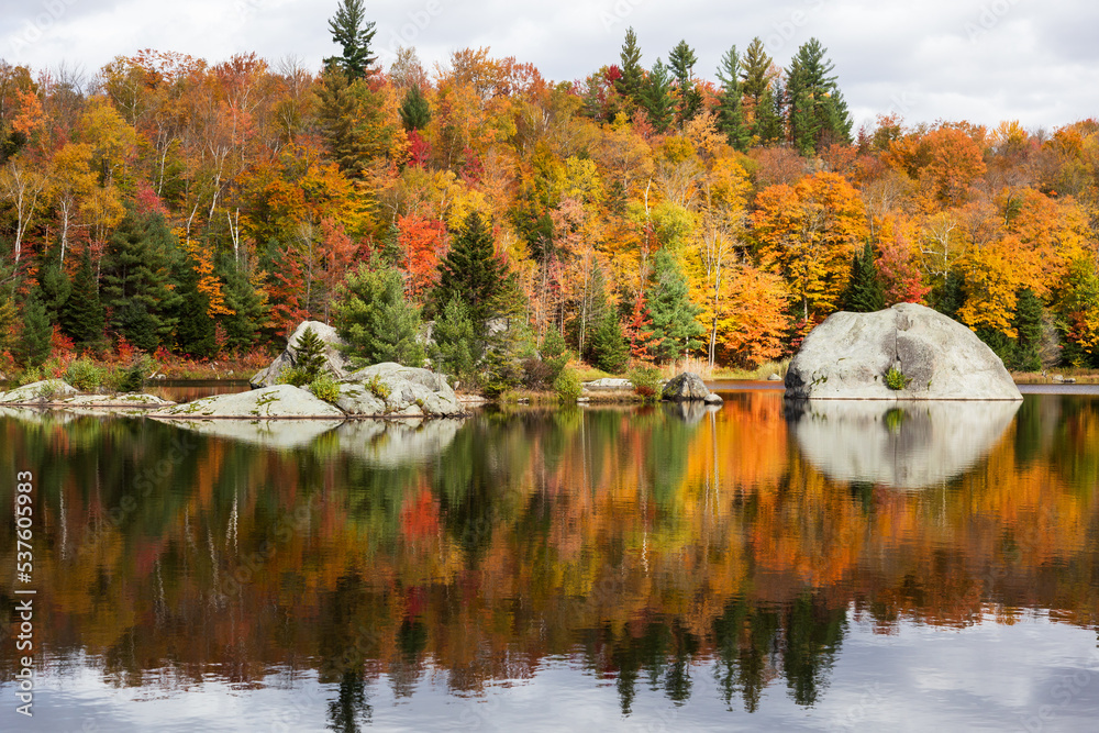 Fototapeta premium Large boulders reflecting in small lake in the Eastern Townships region, with fall foliage in the background, Knowlton, Quebec, Canada