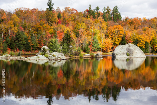  Large boulders reflecting in small lake in the Eastern Townships region, with fall foliage in the background, Knowlton, Quebec, Canada