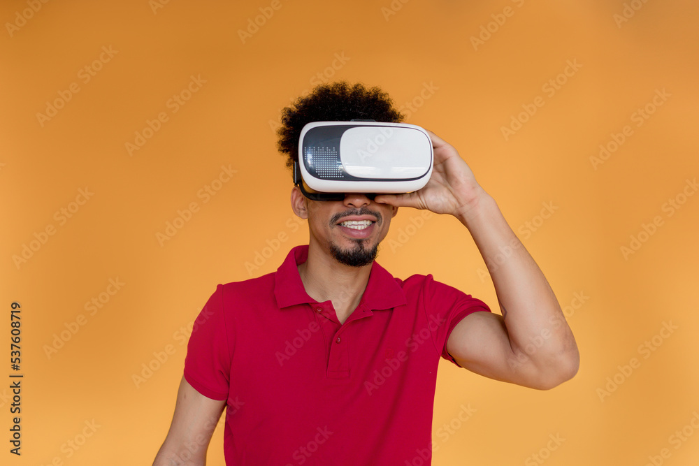 Close up photo of dark-skinned man with wavy dark hair wearing red tee in VR glasses playing in video games over yellow backdrop Close up photo of dark-skinned man with wavy dark hair wearing red tee in VR glasses playing in video games over yellow backdrop