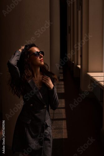 portrait of a woman.  A young attractive woman in sunglasses stands by a pillar of a building and looks out the sunny window.