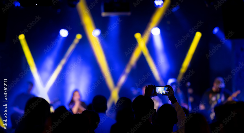 Crowds of people at a live concert show. Rock band on a stage with silhouettes of people hands in the air enjoy the show. Fans filming or taking photos of a live performance. 