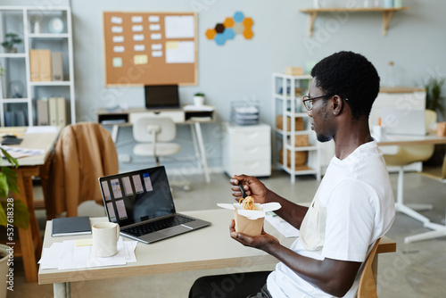 Side view portrait of young black man eating takeout noodles at workplace during lunch break, copy space