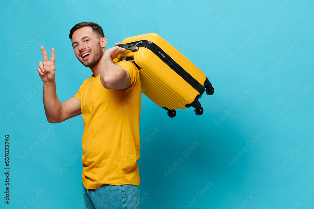 Happy smiling tanned handsome man in yellow t-shirt show v-sign gesture ready for vacation hold suitcase posing isolated on blue studio background. Copy space Banner Mockup. Trip journeys concept