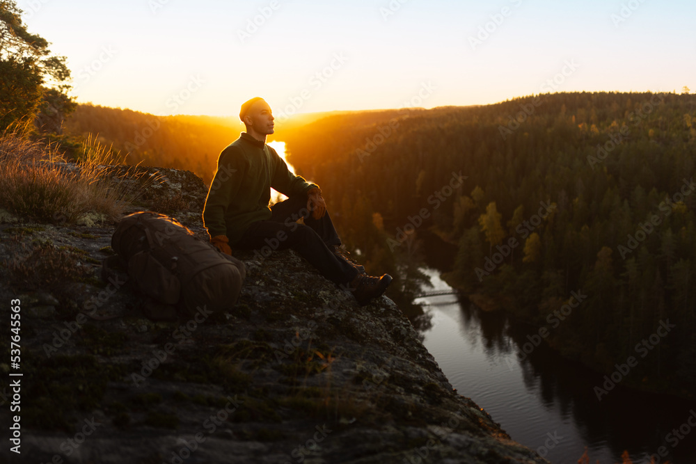 A male hiker wearing a beanie sitting on the top of a mountain at sunrise looking out over a river.