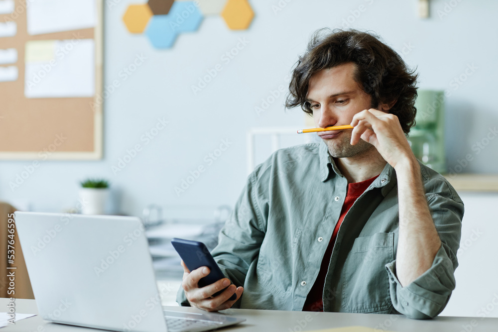 Portrait of Caucasian young man playing with pencil and using phone at workplace suffering from boredom and procrastination - obrazy, fototapety, plakaty 