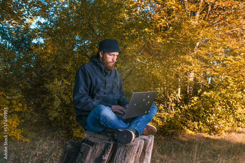 man with a laptop works remotely sitting on a stump in nature
