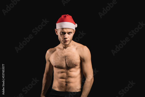 muscular sexy man with a naked torso in a santa claus hat on a dark background. the concept of a gift for the new year and christmas to the athlete and wife. © velimir