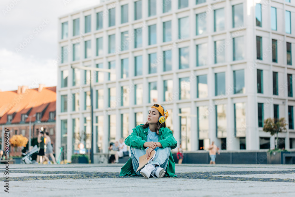 Stylish teenage student girl with headphones sits on city street of Wroclaw, Poland