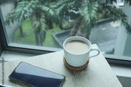 A cup of coffee with a handphone by the window