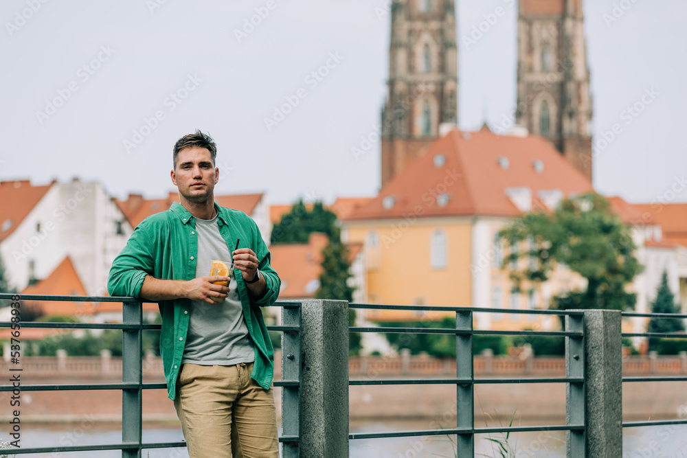 Stylish young man with glass of juice on river bank in Wroclaw, Poland