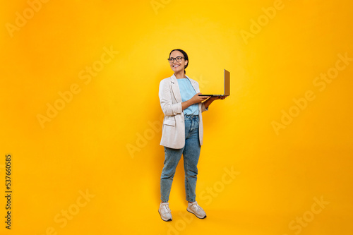 Full length photo of happy successful stylish latino or brazilian young woman, business employee, with glasses, hold in hands open laptop, stand on isolated orange background, looks to the side, smile