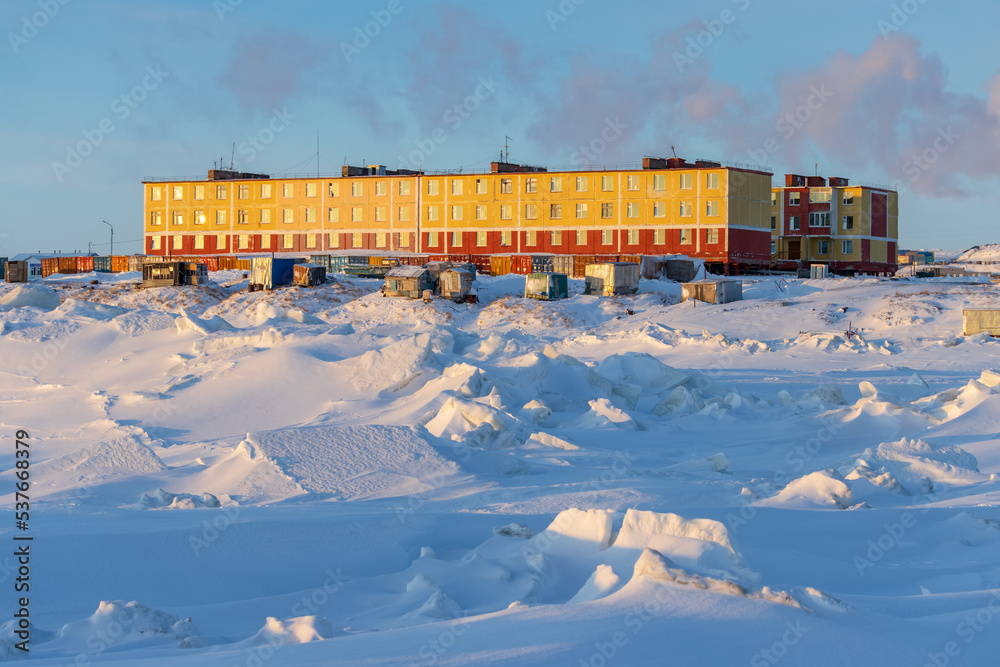 A small northern village in the Arctic. Residential buildings and fishing sheds on the coast of the frozen sea. Cold frosty winter weather. Ice hummocks and snow. Chukotka, the Far North of Russia.