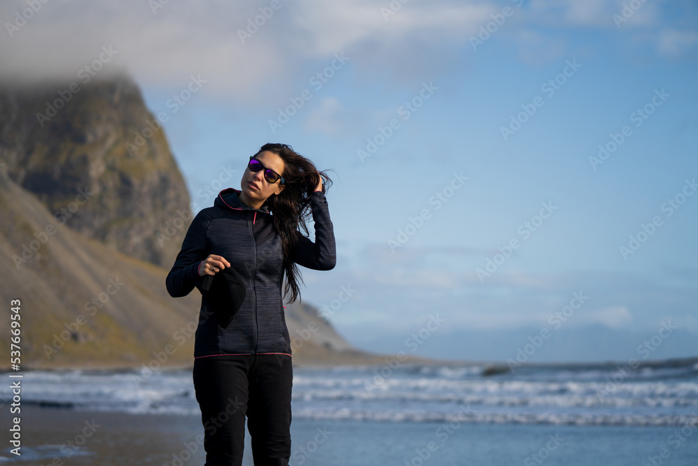 adventurous woman with cap and sunglasses walking on the black volcanic sand beach at the seashore with mountains in autumn winter