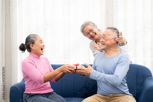 Happy Senior Asian women covering eyes of senior Asian man for surprise with gift box in home. group of happy senior asian giving a gift in living room. happy moment on sofa.
