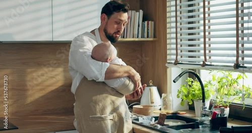 Bearded father in apron holding newbon baby on hands standing near window in modern light kitchen speaking with newborn baby smiling having fun photo