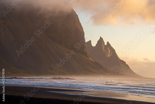 sunrise on a black volcanic sand beach overlooking large mountains and the sea. 